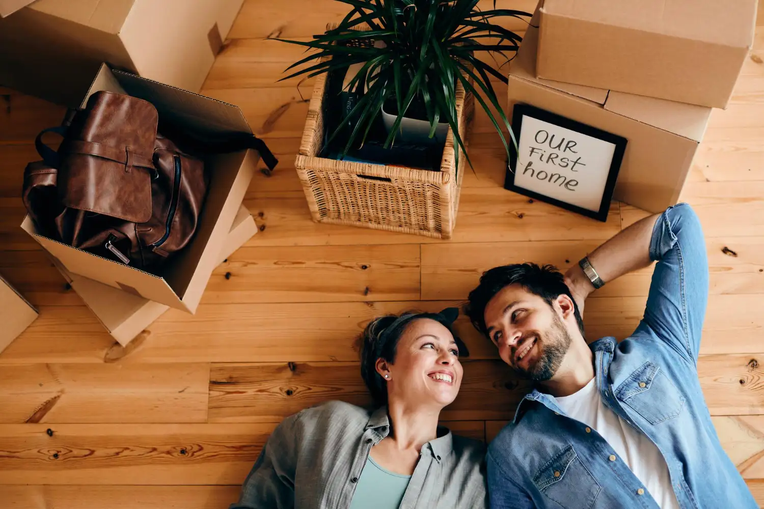 Glückliches Paar liegt zwischen Umzugskartons beim Umzug ins Eigenheim neben einem Schild mit Aufschrift Our First Home