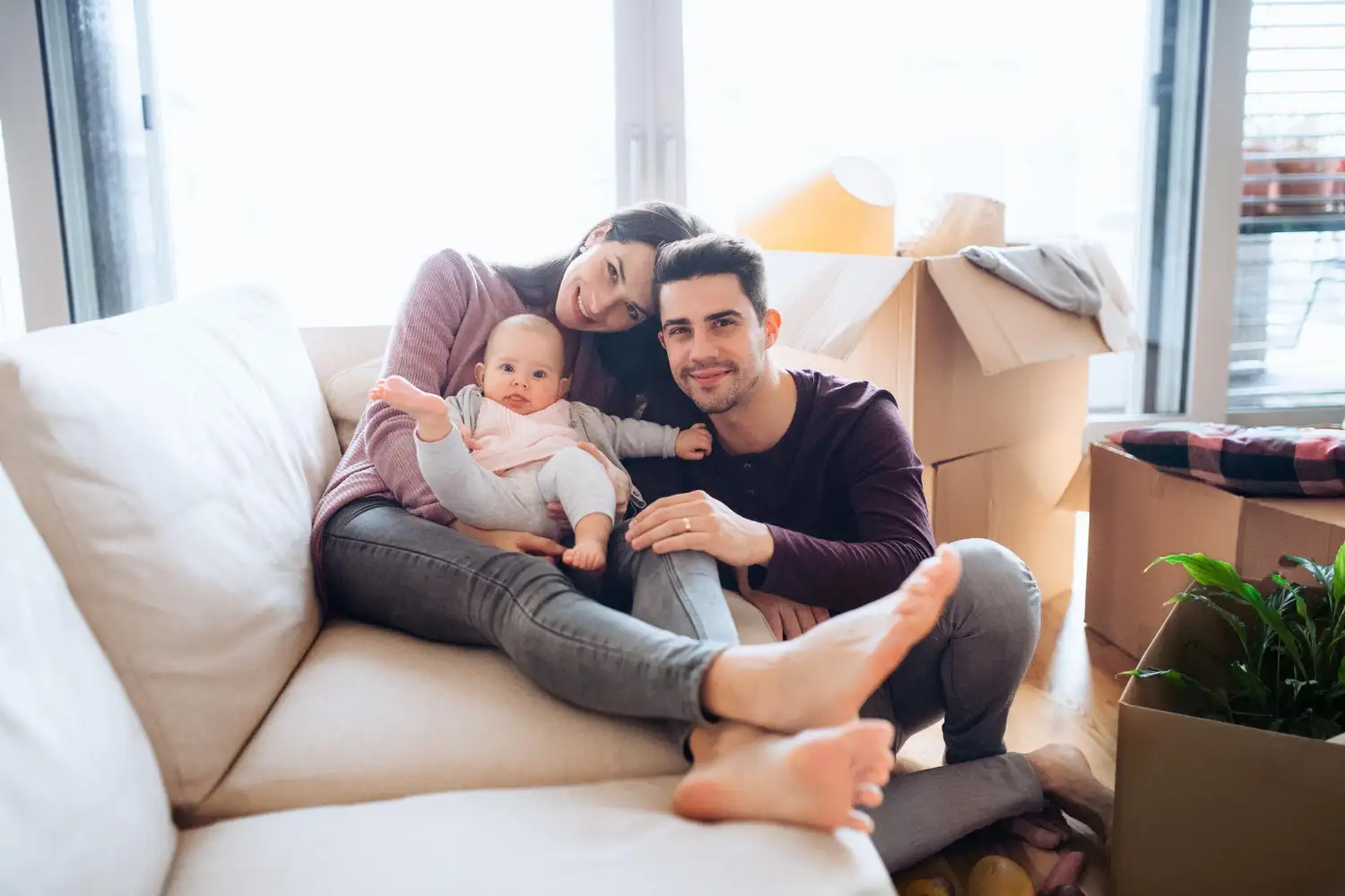 Junge Familie sitzt beim Umzug mit Baby entspannt auf dem Sofa zwischen Umzugskartons in der neuen Wohnung in Wien
